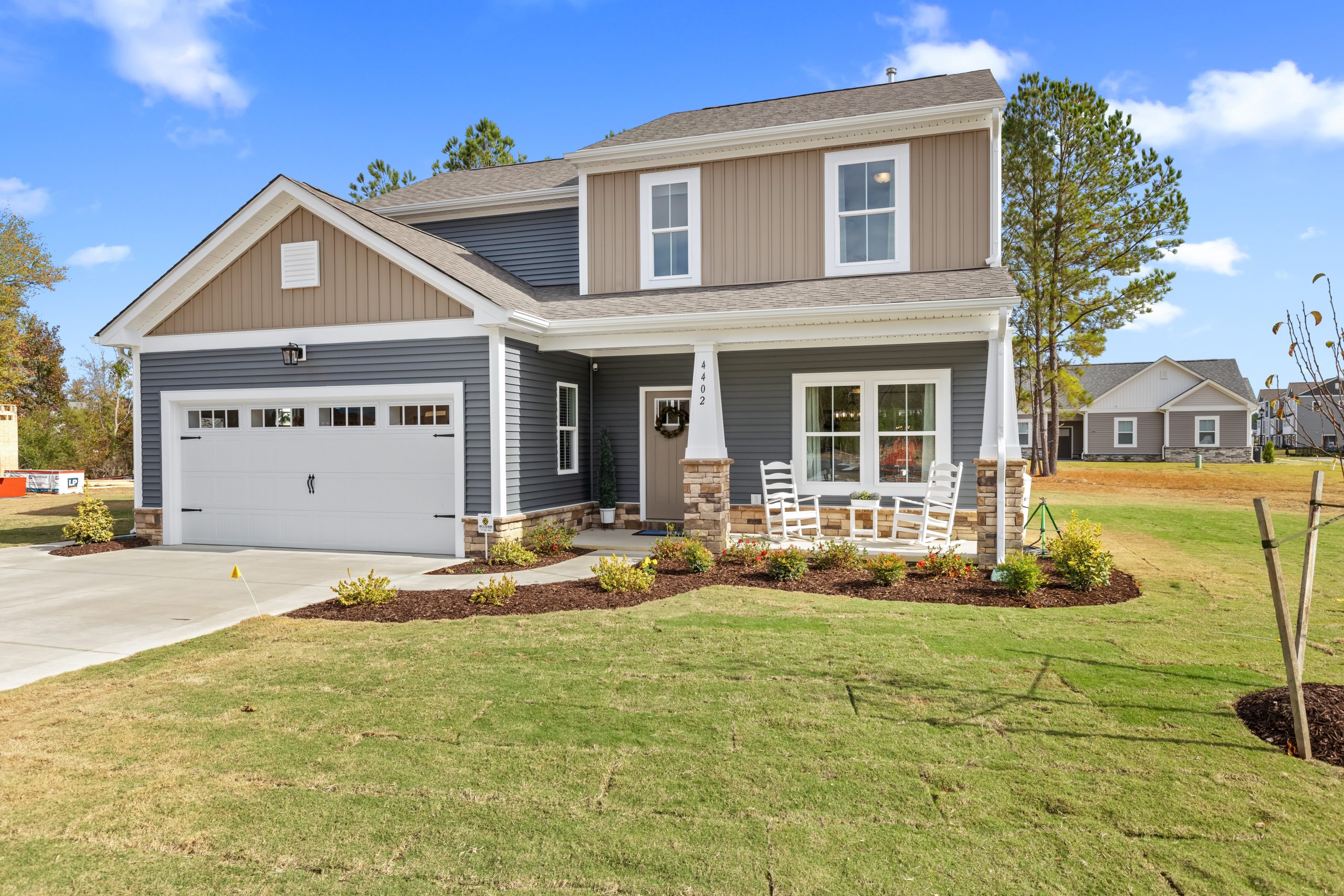 A two-story craftsman-style home with gray and beige siding, white trim, and stone accents on the front porch columns. The house features a two-car garage, covered front porch with white rocking chairs, and professional landscaping with mulched beds and a manicured lawn. The architecture includes craftsman details like white pillars, divided light windows, and board and batten siding accents on the upper level.