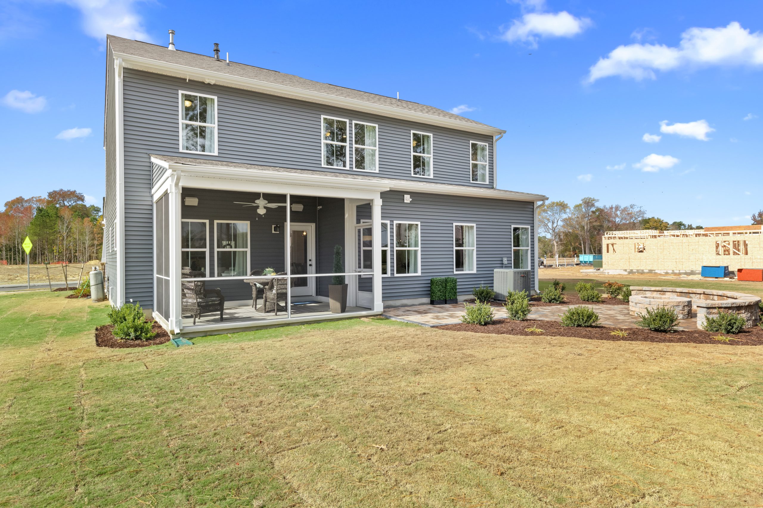 A rear view of a two-story home featuring blue-gray siding with white trim. The back of the house includes a covered porch with screening and outdoor furniture, large windows throughout, and a professionally landscaped yard with mulched beds and stone retaining walls. In the background, new construction can be seen in the developing neighborhood. The home is positioned on a gently sloping lawn with young landscaping.