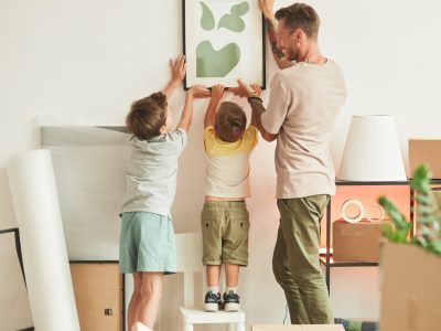 Full length portrait of happy father with two sons hanging pictures on wall while moving in to new home